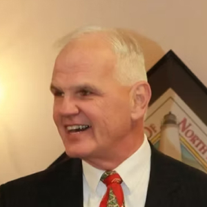 A smiling older man with short gray hair, wearing a dark suit, white shirt, and red patterned tie, stands in front of a beige wall with a framed picture in the background.