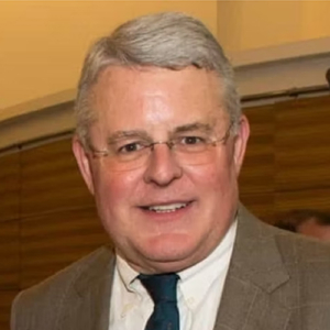 A middle-aged man with short gray hair and glasses is smiling. He is wearing a brown plaid suit jacket, a white shirt, and a dark tie. The background is indoors with warm lighting and blurred details.