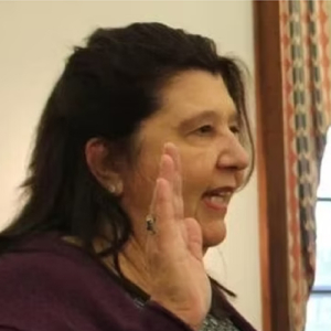 A woman with dark hair raises her right hand, as if taking an oath. She is indoors, with patterned curtains and a window visible in the background.
