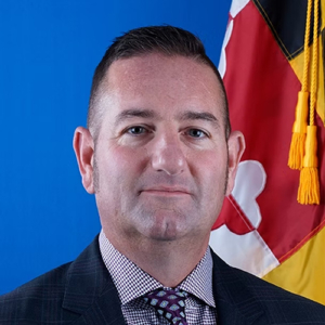 A man in a checkered dress shirt, patterned tie, and dark jacket poses in front of a blue background and part of a flag with red, white, black, and gold colors.