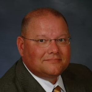 Portrait of a man with short hair, wearing glasses, a gray suit, white shirt, and yellow tie, set against a dark blue background.