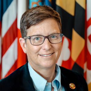 Professional headshot of Sandra, a smiling woman with glasses, against a Maryland state flag backdrop. Portrait photography.